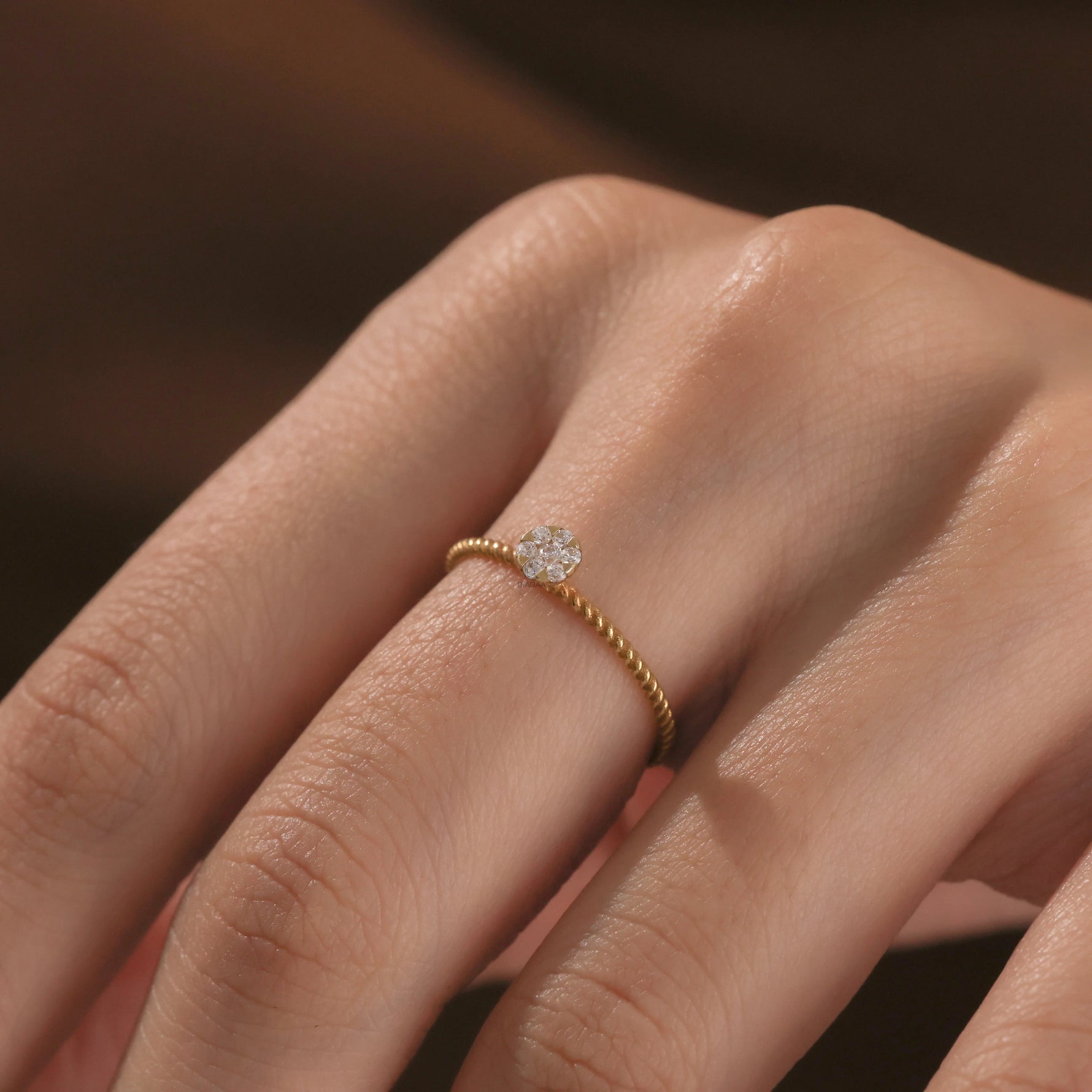 Detailed view of a fine Close-up of a hand wearing a dainty yellow gold twisted band ring featuring a round small diamond cluster centerpiece.
