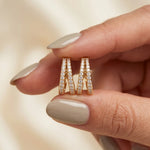 A hand with glossy taupe manicured nails holding a pair of yellow gold V-shaped split hoop earrings encrusted with double rows of pavé diamonds.