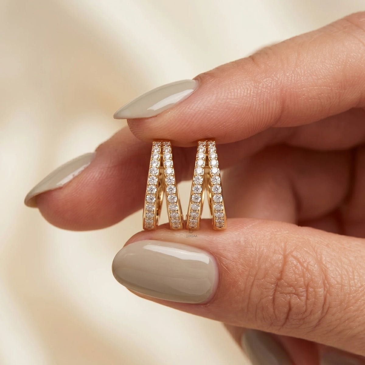 A hand with glossy taupe manicured nails holding a pair of yellow gold V-shaped split hoop earrings encrusted with double rows of pavé diamonds.