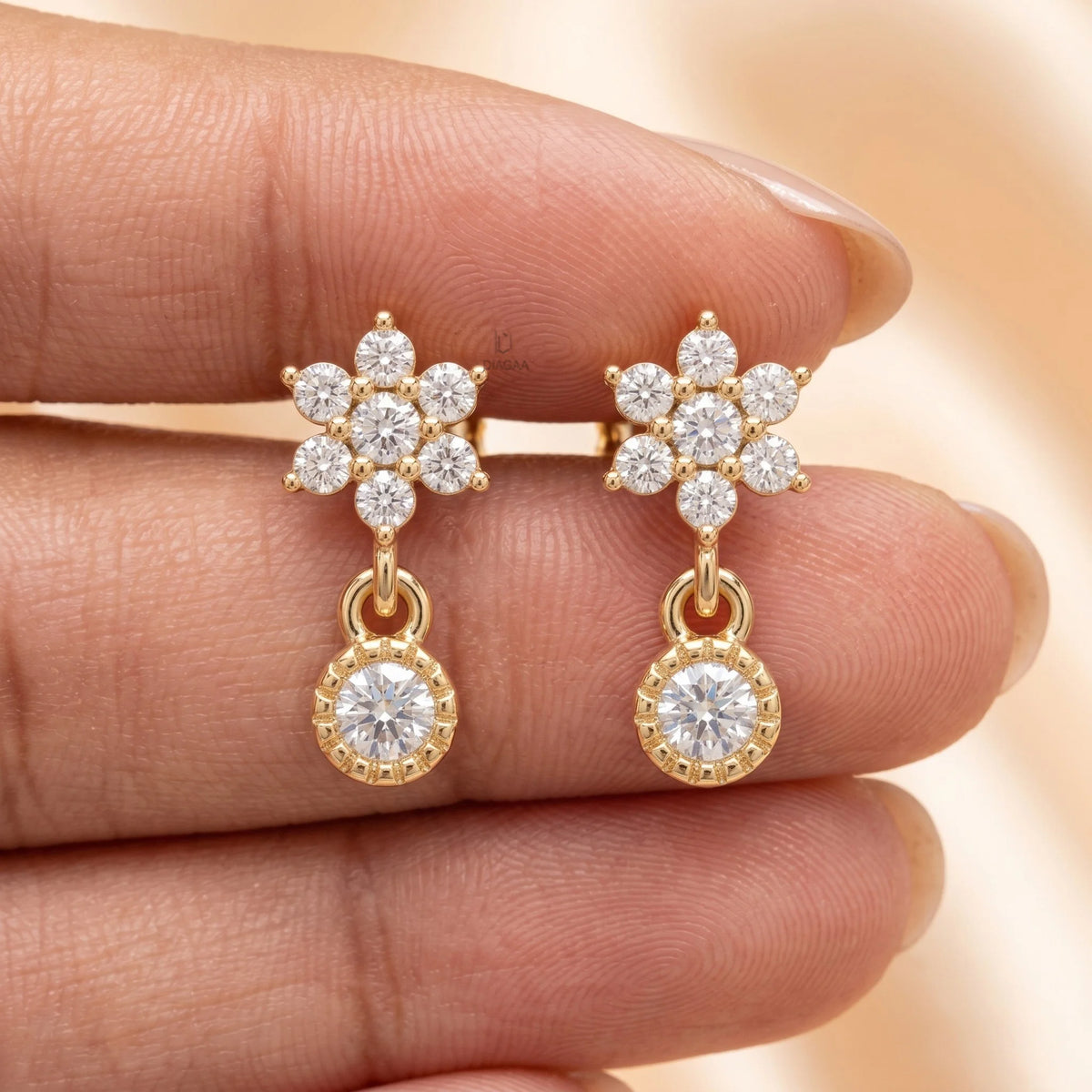 Hand holding a pair of yellow gold floral diamond drop earrings featuring a flower cluster stud and dangling round stone.