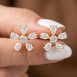 Close-up shot of a prong-set pear and round diamond stud earring pair held between fingers. Designed in solid yellow gold with a five-petal floral cluster setting.
