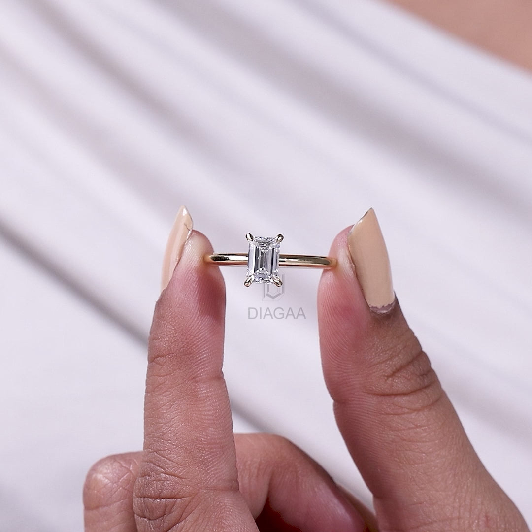 Solitaire emerald-cut diamond engagement ring in yellow gold held up and worn on a manicured hand.