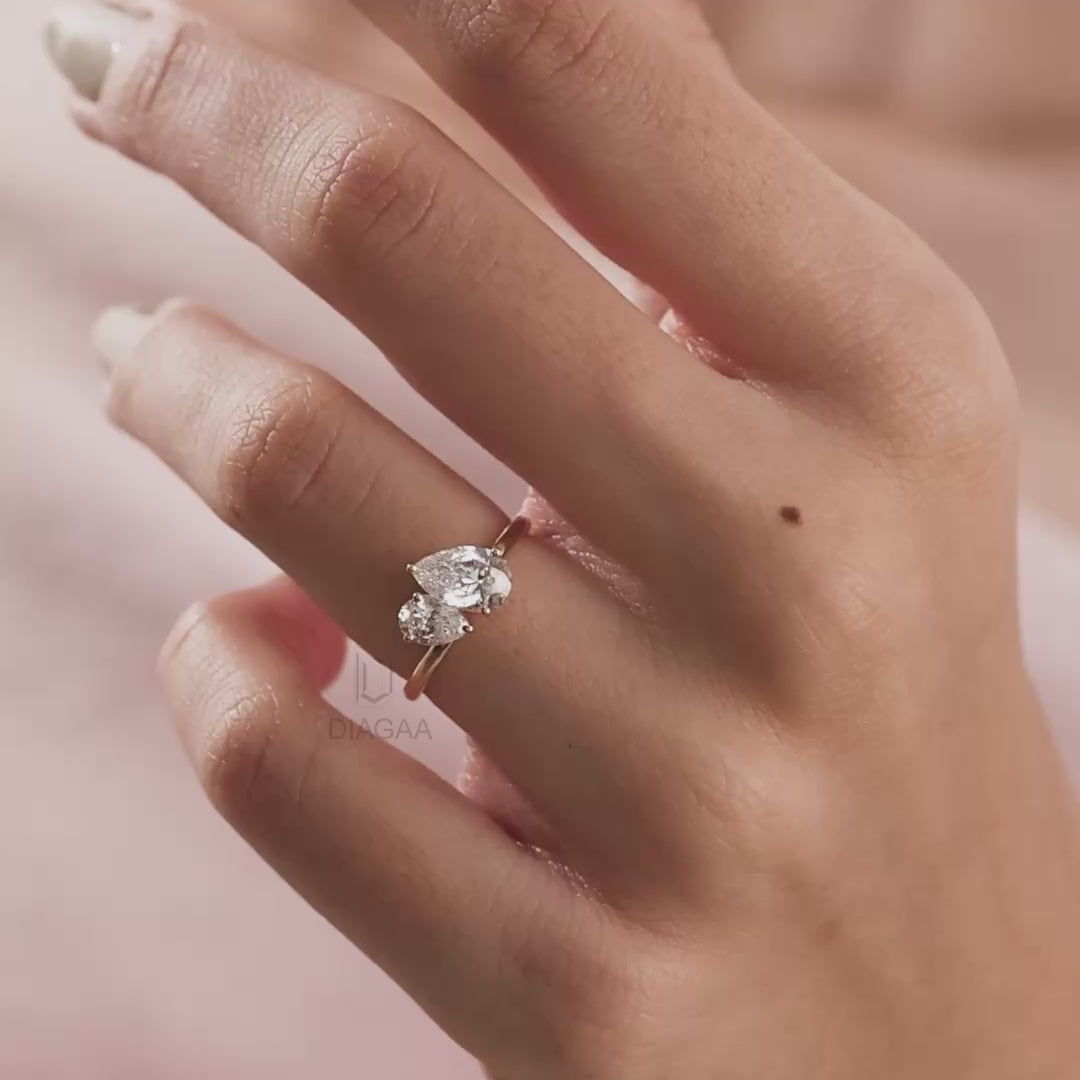 Stunning Toi et Moi gold engagement ring featuring two pear-cut diamonds worn on a finger against a soft pink background.