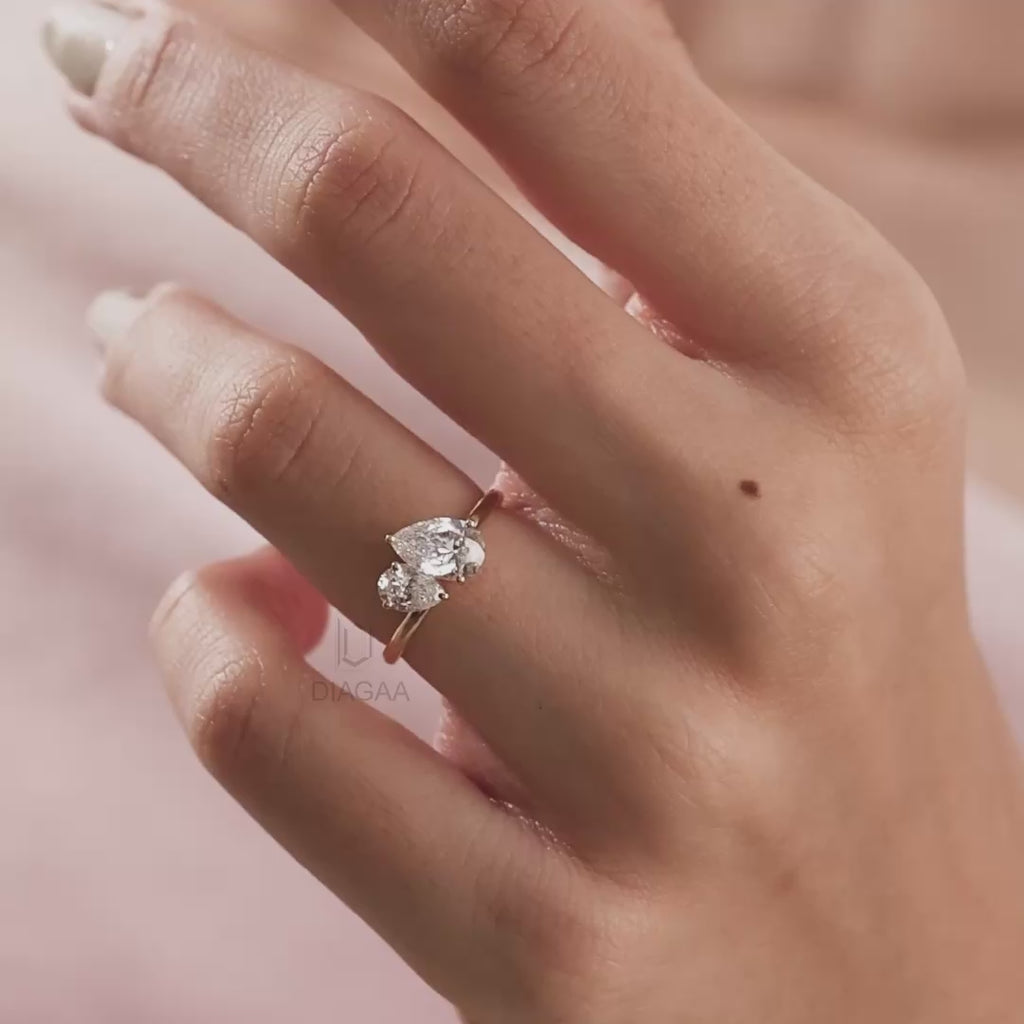 Stunning Toi et Moi gold engagement ring featuring two pear-cut diamonds worn on a finger against a soft pink background.