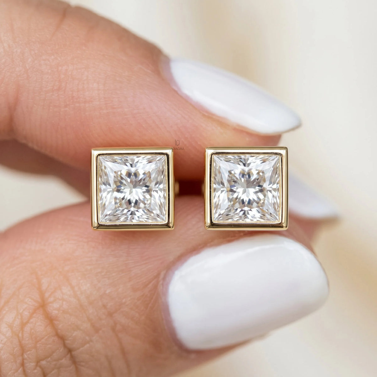Hand holding classic square princess-cut diamond stud earrings set in smooth yellow gold full bezels earring.