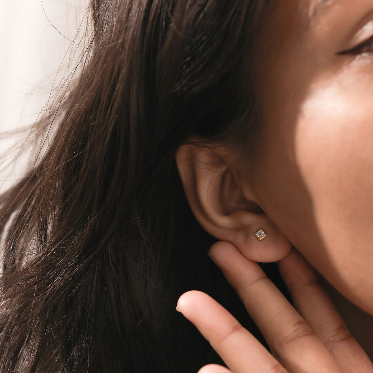 Close-up of a woman wearing a minimalist gold square stud earring with a diamond center. Dainty everyday jewelry on an earlobe piercing.