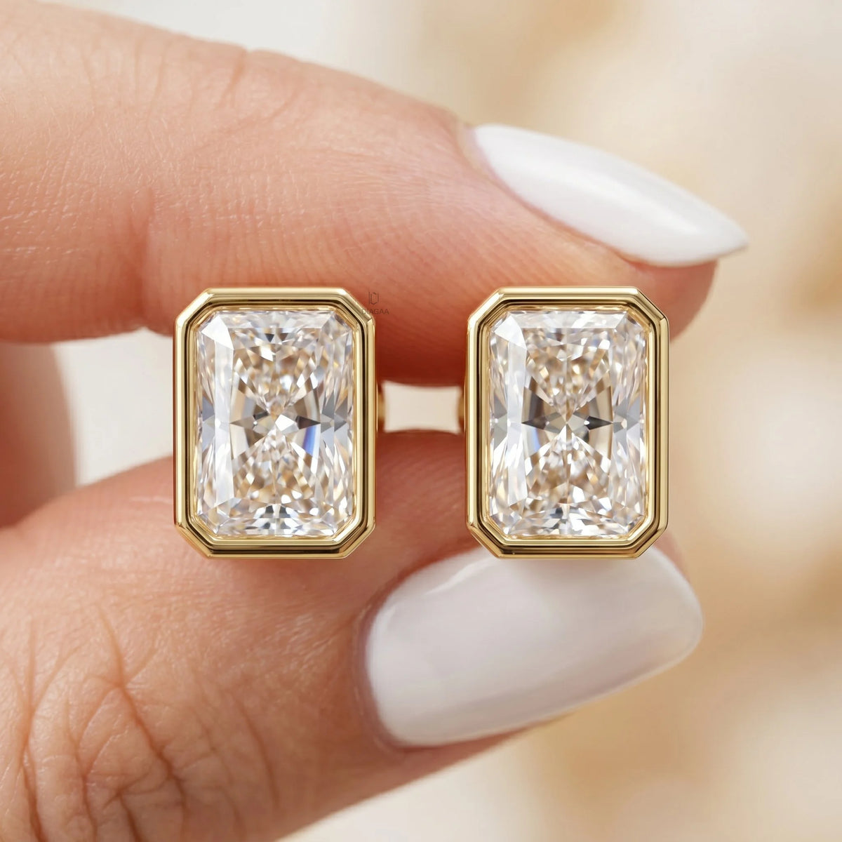 A close-up of a person holding a pair of radiant-cut diamond stud earrings featuring a modern yellow gold bezel setting earring.